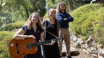 Vor grünen Hecken stehen zwei junge Frauen, eine mit Gitarre, und ein junger Mann