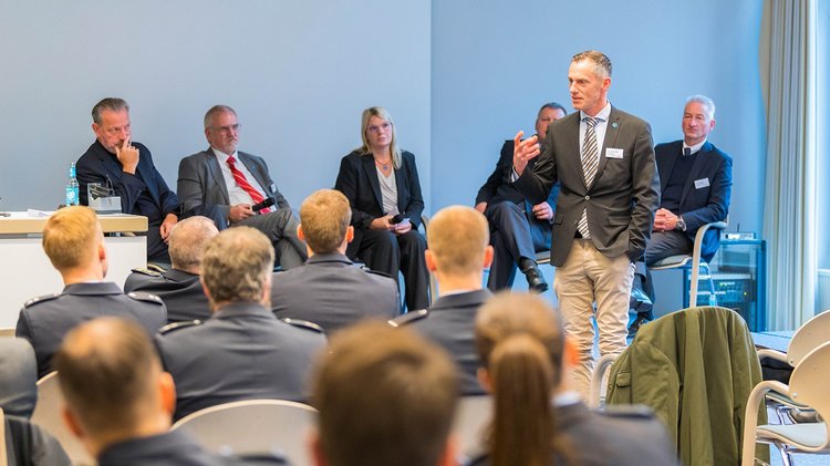 Sechs Menschen sitzen auf Podium, einer steht und spricht, vorn Publikum