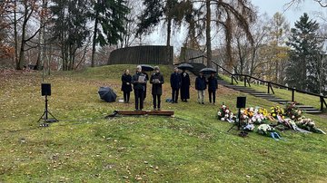 Kleine Menschengruppe mit Regenschirmen, rechts Gedenkkränze