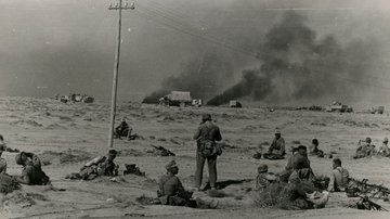 Soldaten sitzen im Sand. Im Hintergrund befinden sich brennende Fahrzeuge.