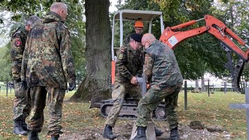 Männer in Uniform setzen Grabkreuze, im Hintergrund steht ein Bagger.