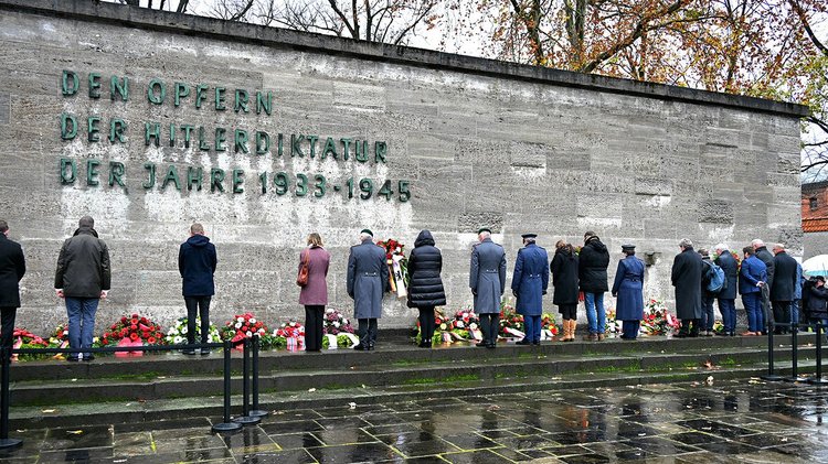 Menschen in einer Reihe von hinten vor Kränzen am Fuß einer hohen Mauer