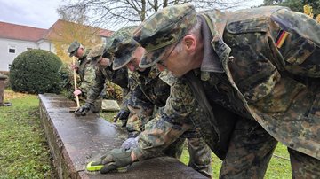 Männer in Uniform reinigen eine Steinmauer auf einem Friedhof.
