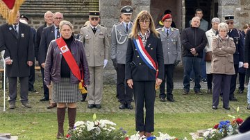 Zwei Frauen stehen schweigend vor Kränzen am Boden, hinten Publikum