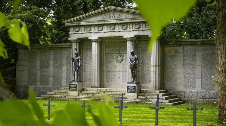 Blick unter Bäumen hindurch auf zwei bronzene Männerstatuen vor steinernem Portal