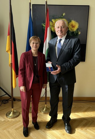 Frau und Mann, der eine Auszeichnung zeigt, im Hintergrund Fahnen