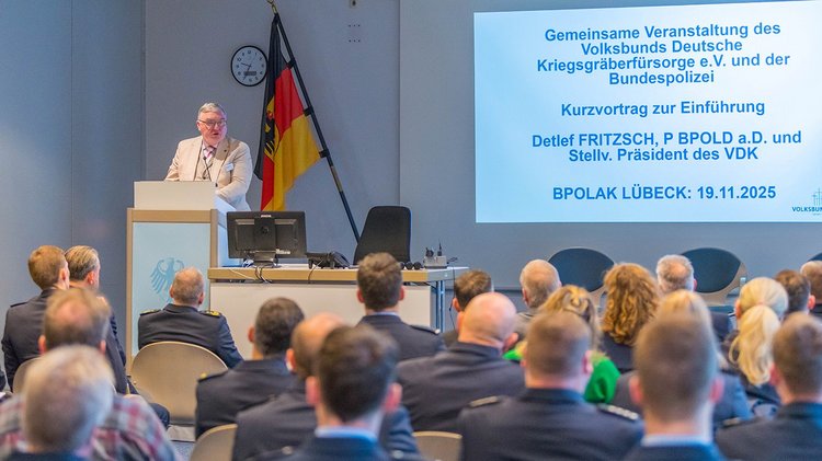 Mann am Rednerpult vor Deutschlandfahne und Bildschirm, vorn Publikum