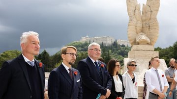 Im Vordergrund: eine Reihe festlich gekleideter Menschen vor einem Denkmal. Im Hintergrund: ein Kloster auf einem Berg