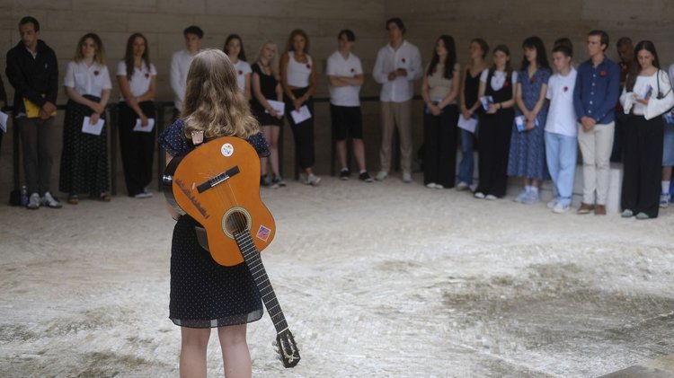 Im Hintergrund: Jugendliche in einer Reihe. Im Vordergrund: die Rückenansicht einer jungen Frau mit Gitarre