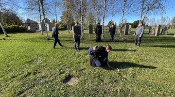 Frau legt auf weitläufigem Friedhofsgelände Blume auf dem Rasen ab, im Hintergrund mehrere Menschen