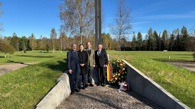 Drei Männer und eine Frau mit Gedenkkränzen auf Friedhof in betonierter, in den Boden eingelassener Einfassung des Hochkreuzes