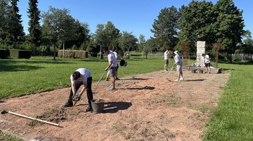 Jugendilche reinigen Sandfläche vor Denkmal mit Harken und Hacken
