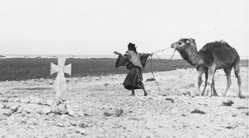 In einer Steinwüste sieht man ein Grabkreuz, daneben einen Beduinen mit einem Kamel.