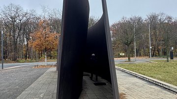 Mensch geht durch ein begehbares Kunstwerk mit zwei hohen, geschwungenen Wänden aus dunkelgrauem Stahl
