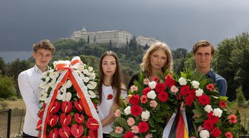 Im Vordergrund: vier Jugendliche - zwei Jungen, zwei Mädchen - die Kränze halten, einen mit polnischer, einen mit deutscher Schleife. Im Hintergrund: das Kloster Monte Cassino auf einem Berg.