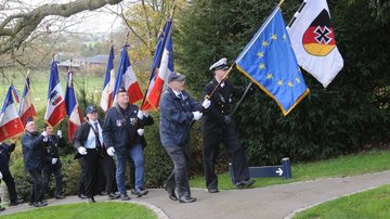 Verschiedene Uniformierte erklimmen von links nach rechts aufwärts einen Hügel. Sie tragen Fahnen der Bundeswehr, der Europäischen Union und der französischen Republik.