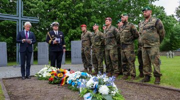Vier Blumengestecke auf dem Boden, eine Gruppe Soldaten und zwei Redner vor einem Hochkreuz 