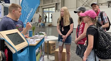 Informationsstand in der Messehalle: Junger Mann erklärt drei Jugendlichen und einem Erwachsenen den Volksbund.