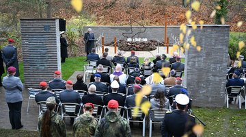 Stuhlreihen mit Publikum von hinten zwischen Steinstelen und vor Mahnmal