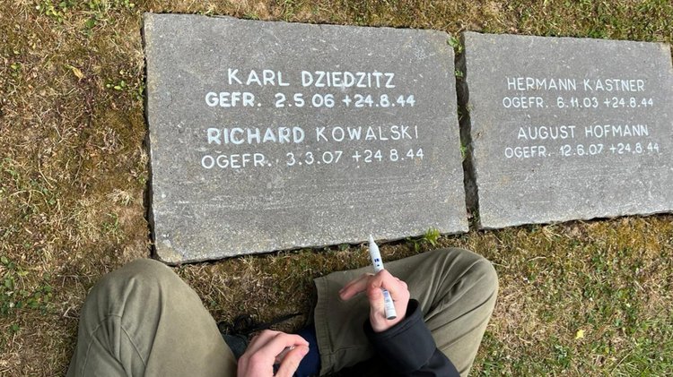 Hand mit Edding über Grabplatte mit weißer Inschrift